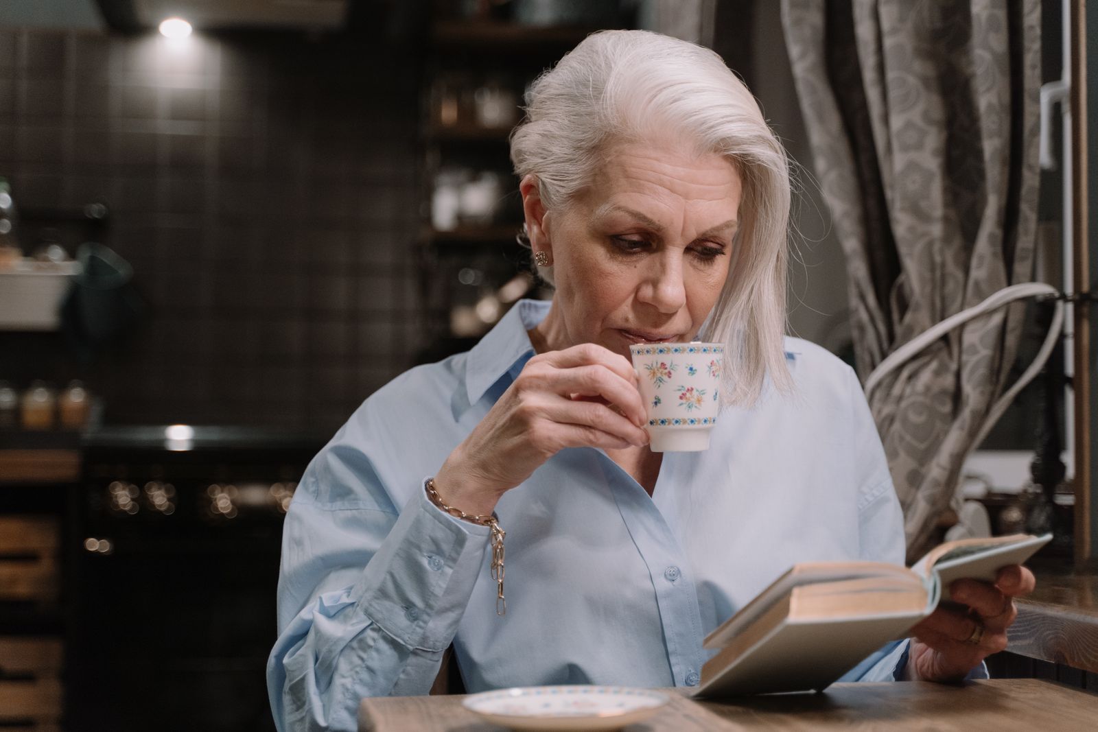 An older woman with white hair sipping tea from a floral cup while reading a book in a warm home kitchen.