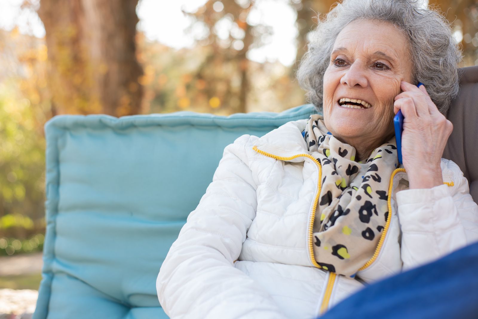 An older woman smiling while talking on a mobile phone, sitting outdoors on a cushioned bench.