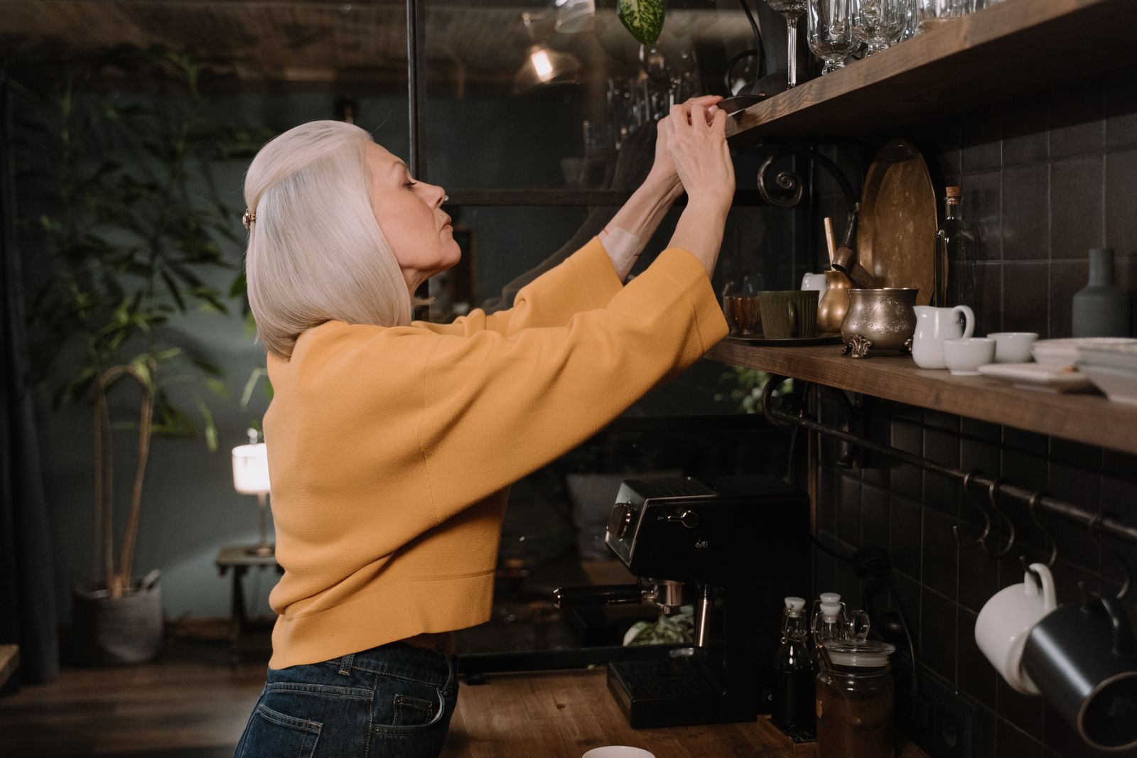 An older woman reaching up to a high kitchen shelf.